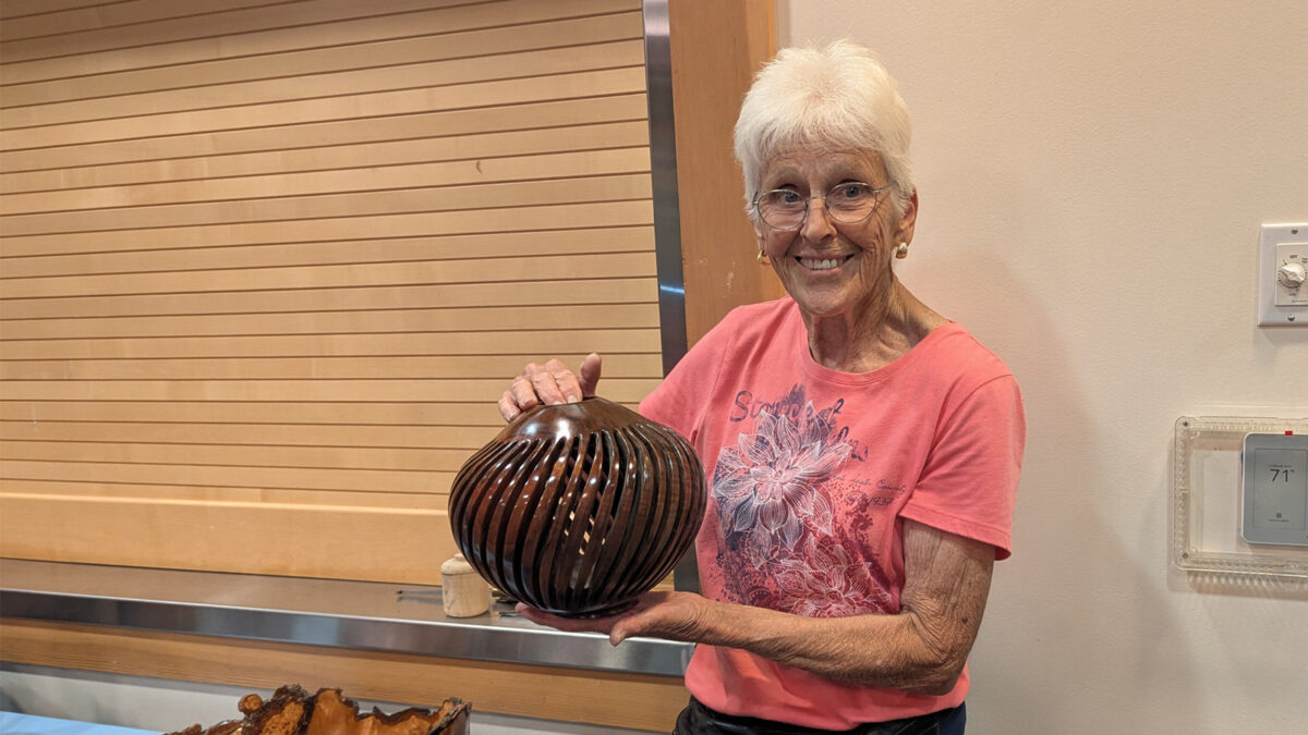 Dottie Bang holds her dark wood (walnut) spiral vase in front of a basswood slat wall.