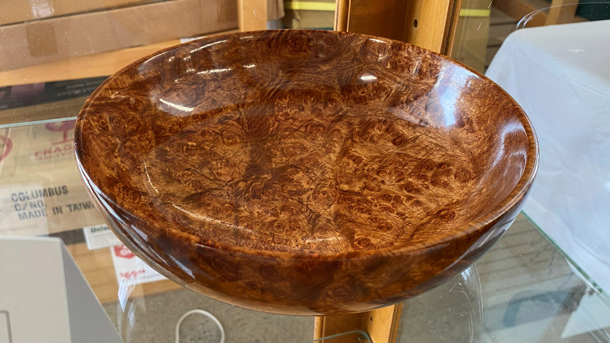 A close-up, high-angle shot of a polished wooden bowl made from Amboyna burl on display at the BAWA woodworking show. The wood features a rich, reddish-brown hue with a complex, swirling grain pattern filled with small eyes and knots. The bowl is wide and shallow with a thick, rounded rim and a high-gloss finish that reflects the overhead lighting. Ed Kaplan created this piece.