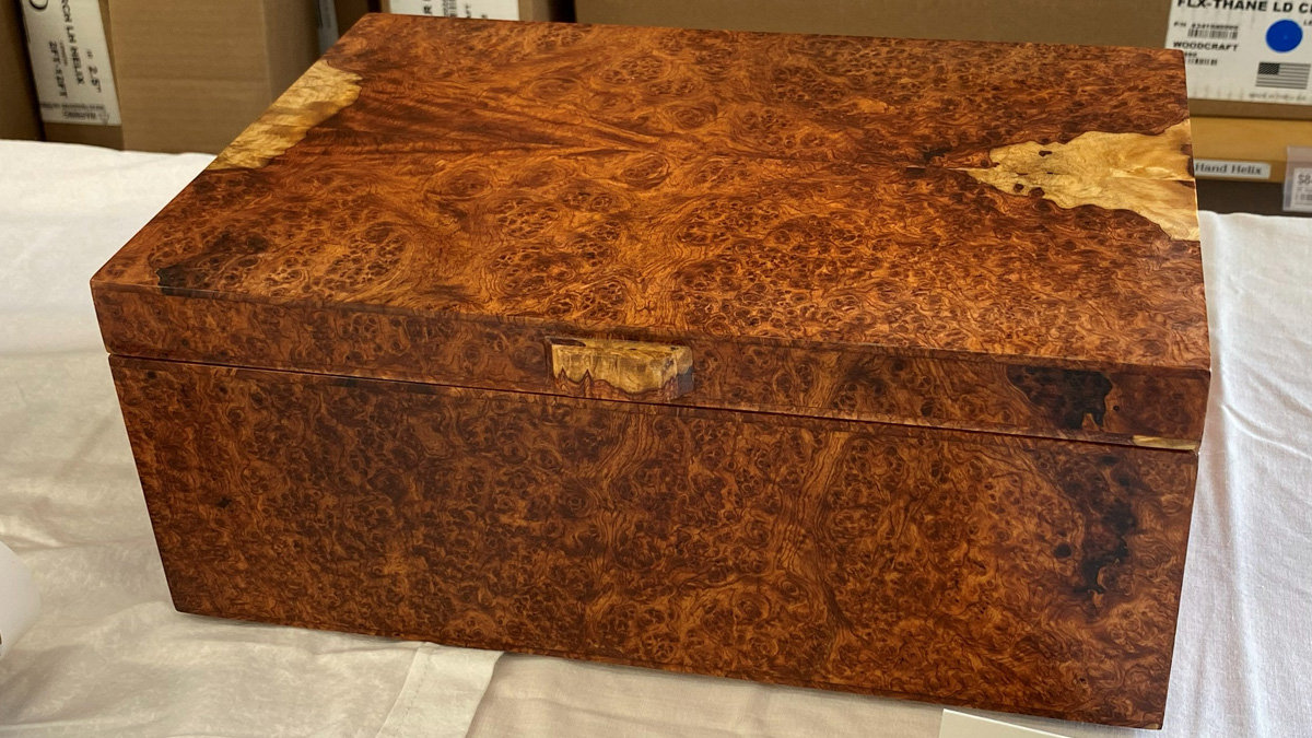 A rectangular wooden lidded box made of highly figured Amboyna burl displayed at the BAWA woodworking show in 2026. The wood features a rich, reddish-brown hue with complex swirling grain and small "eyes" throughout. There are patches of lighter, pale yellow wood on the top surface and a small wooden latch centered on the front edge of the lid.