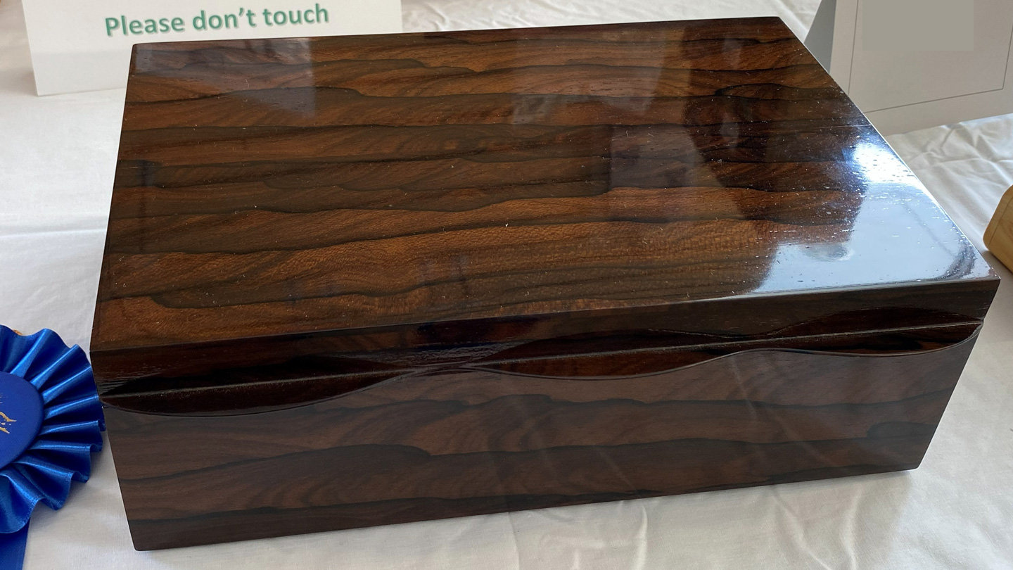 A rectangular wooden box crafted from dark Zircote wood, featuring a high-gloss finish that reflects the light. The wood has distinctive, wavy grain patterns running horizontally across the lid and sides in shades of deep brown. The lid and base are separated by a subtle, undulating curve that creates a gentle wave effect along the edges. A blue prize ribbon is partially visible on the left side.