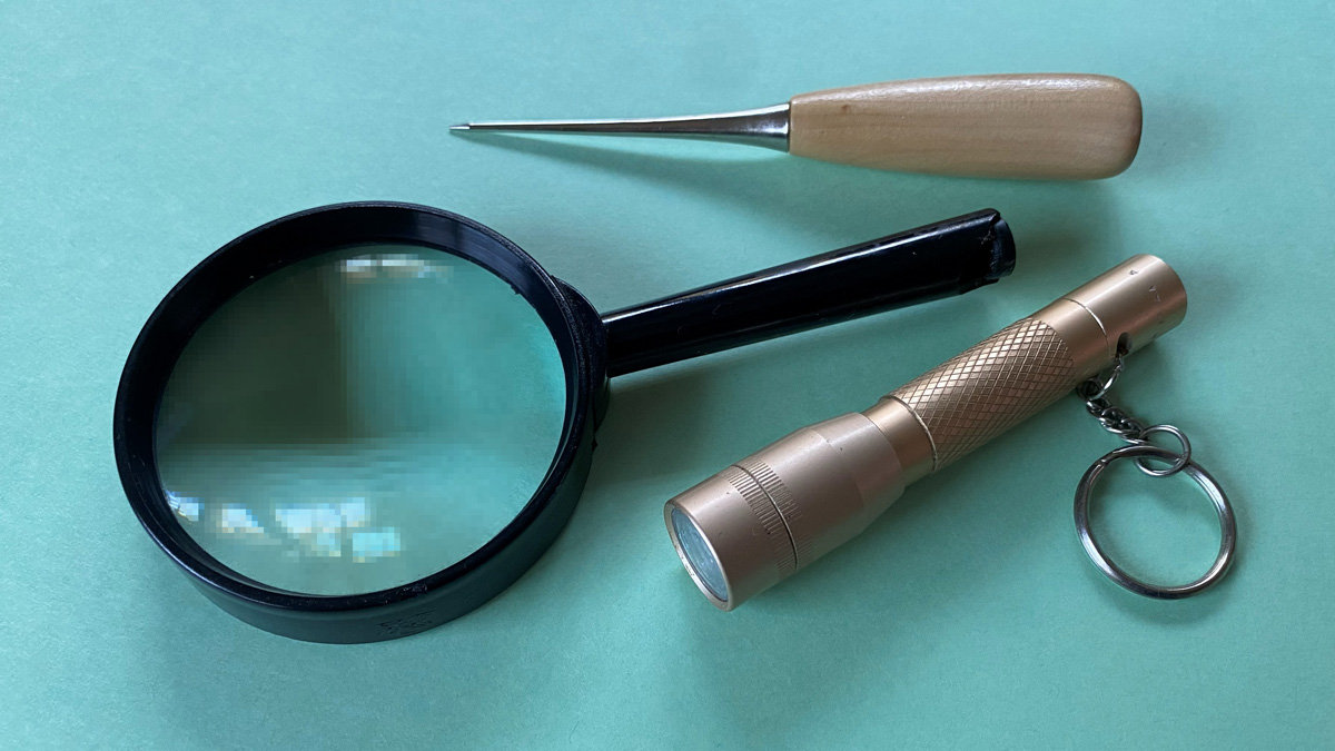 A collection of useful tools arranged on a teal background: a wooden-handled awl at the top, a black magnifying glass in the center, and a brass keychain flashlight below.