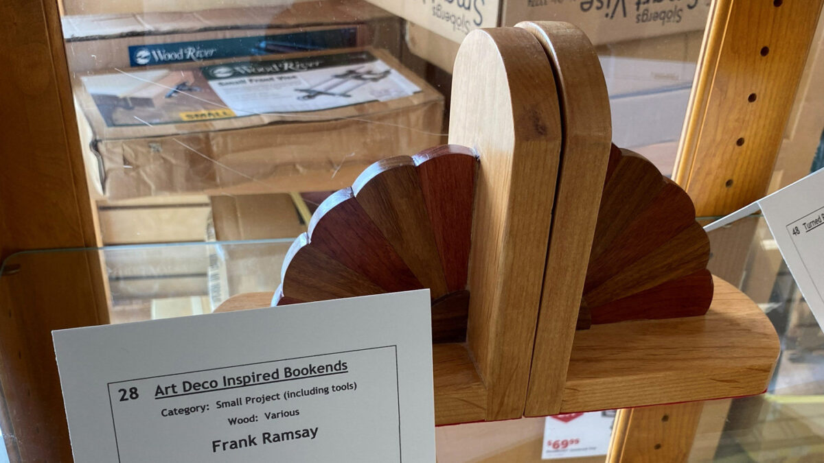 A pair of Art Deco-inspired wooden bookends displayed on a glass shelf at a woodworking show organized by the Bay Area Woodworking Association (BAWA). The bookends feature a light-colored wood frame with a decorative, semi-circular fan design on the side made of alternating segments of dark and reddish-brown wood. A white card in the foreground identifies the project as number 28, "Art Deco Inspired Bookends" by Frank Ramsay, made from various woods.