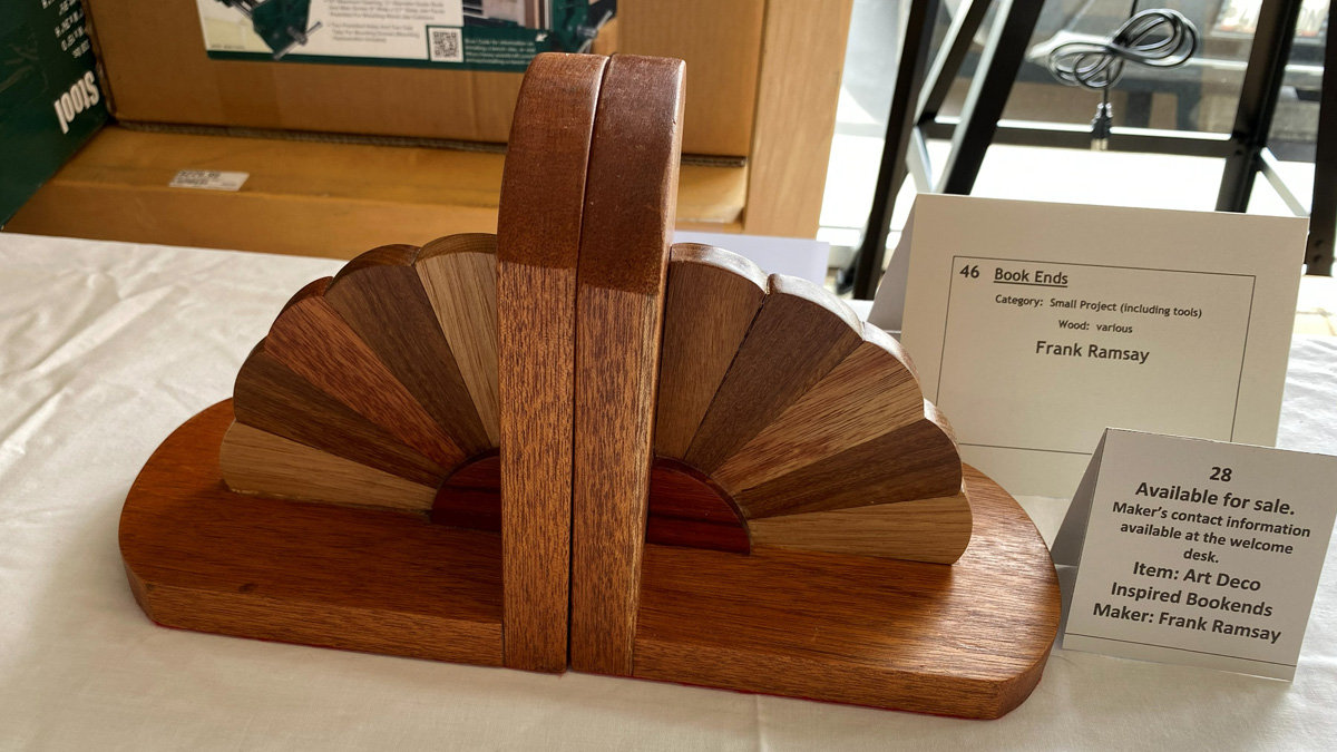 A wooden bookend designed in an Art Deco style by maker Frank Ramsay. The piece features a long, curved base supporting a central vertical divider. On either side of the divider, fan-shaped wings are constructed from alternating radial wedges of light and dark woods, creating a textured, multi-toned aesthetic. Informational cards next to the object identify it as "Art Deco Inspired Bookends" made from various woods.