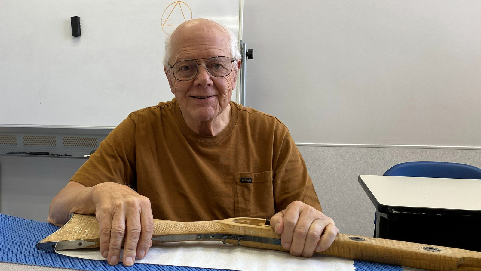 Gary Paulson, a member of the SCVC woodcarving club, sits at a table smiling, holding a long, intricately carved wooden r. stock across his lap; he wears glasses and a brown t-shirt, while the light-colored wood features detailed relief carving along the side panel.