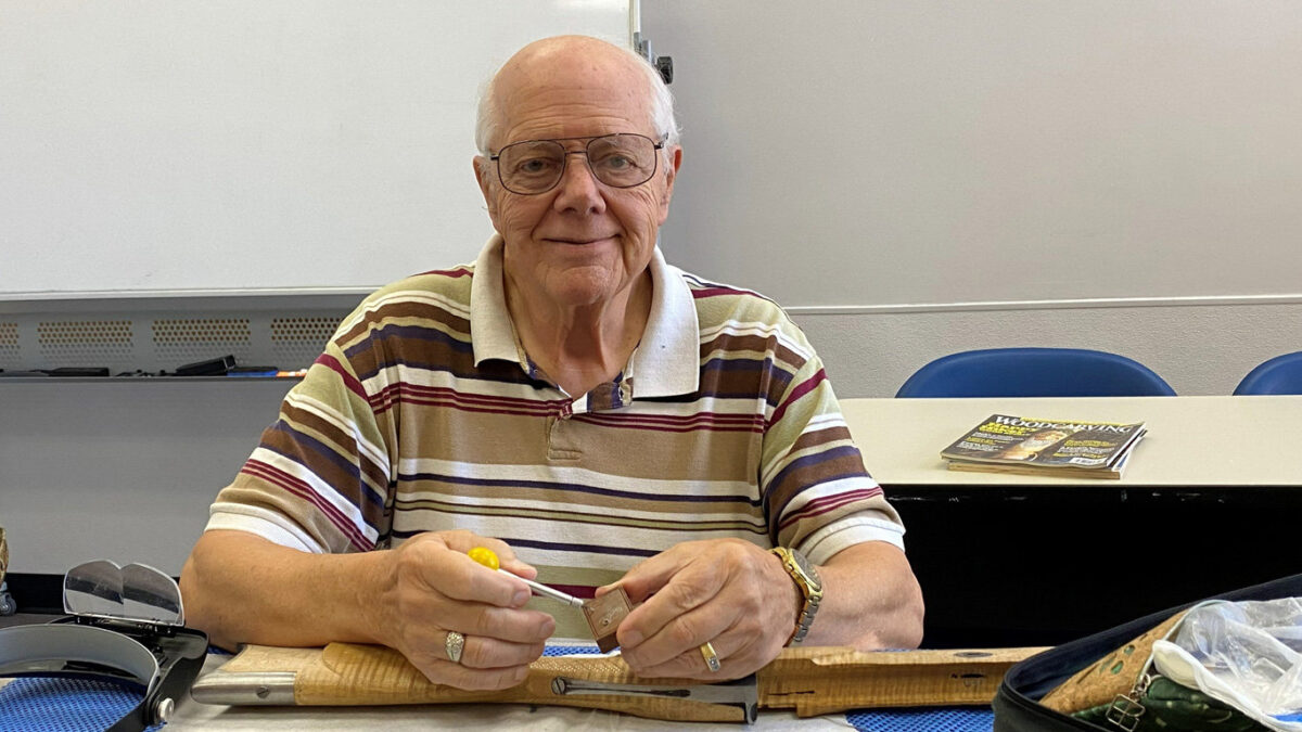 A man, Gary Paulson, sits at a table holding a carving chisel and a small piece of walnut.