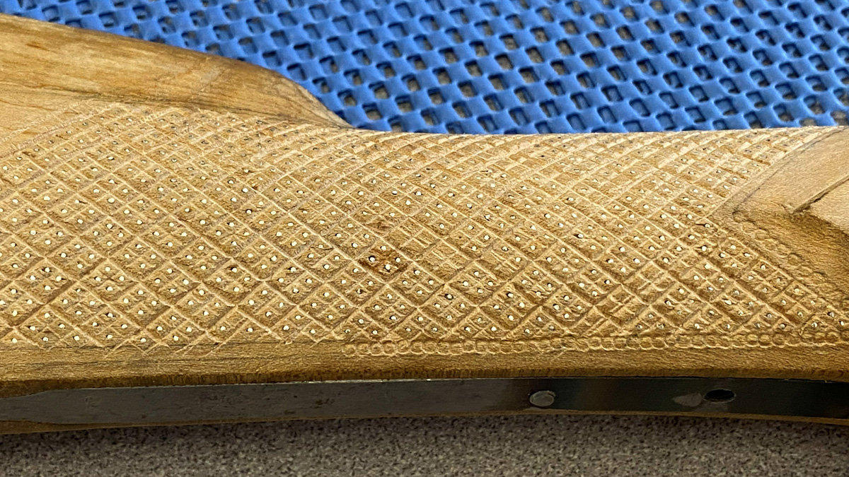 A close-up view of a light-colored wooden item featuring intricate diamond checkering. The pattern is accented by tiny, regular silver inlays placed at intervals within the diamonds. Along the bottom edge, a dark metal bar with screws is visible against a blue perforated background.