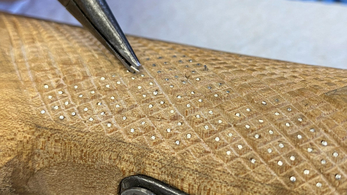 Close-up view of a wooden surface featuring an intricate checkering pattern. The wood has been carved into a grid of raised diamonds, each containing a small, precise silver dot in its center. A pair of fine-point metal pliers is visible at the top left, holding a small silver nail to be hammered into a delicate carving. At the very bottom edge, a curved piece of dark metal hardware is partially visible.