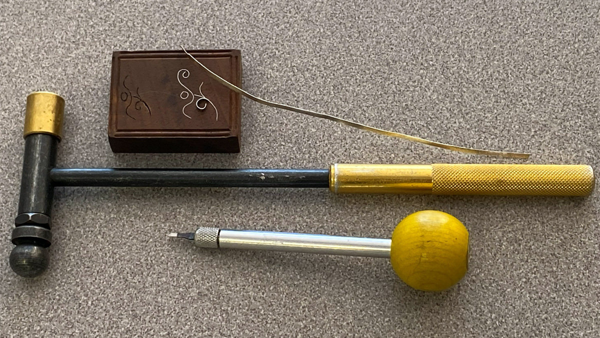 Silver wire inlay tools, including a specialized hammer and graver, are displayed alongside a loose strip of silver wire and a wooden block featuring finished scrollwork designs.