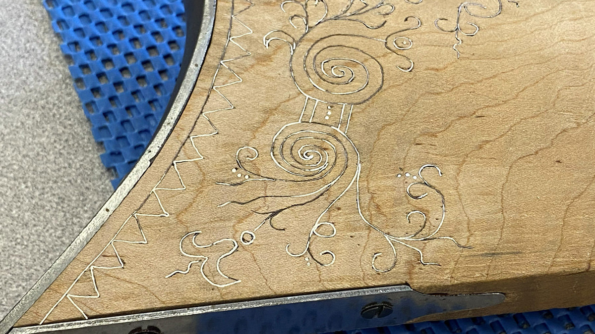 A close-up view of a light-colored wooden surface with intricate silver wire inlay. The central design features two large spirals connected by a rectangular band detailed with small dots, surrounded by delicate, swirling vine-like tendrils and scrolls. A geometric zig-zag pattern runs along the curved left edge, while a metal bracket is visible attached to the bottom edge of the wood.