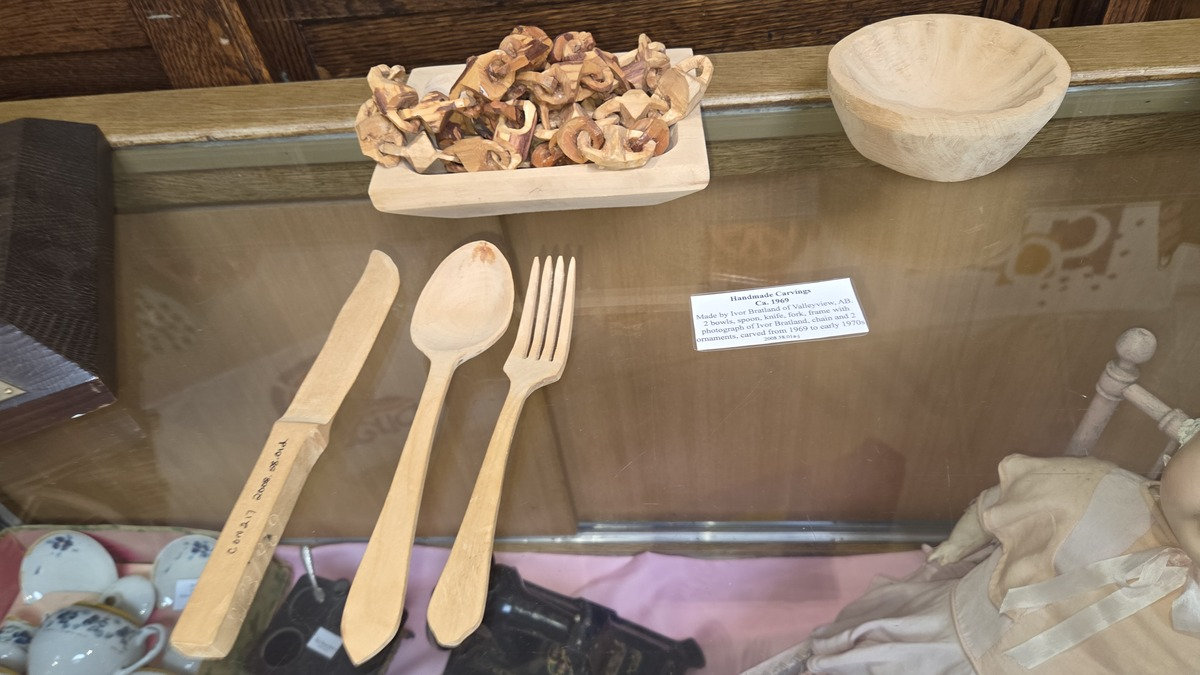 Handmade wooden utensils, including a knife, spoon, and fork, and a light-colored turned bowl carved by Ivor Bratland, displayed in a museum in Drumheller, Canada.