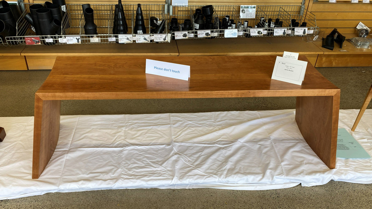A minimalist cherry wood bench with a flat rectangular top and distinctive legs that curve outward and sweep down to the floor. The bench is displayed on a white drop cloth, with a small card on its surface: a project label identifying the piece as a "Bench" by Jamie Baxton.
