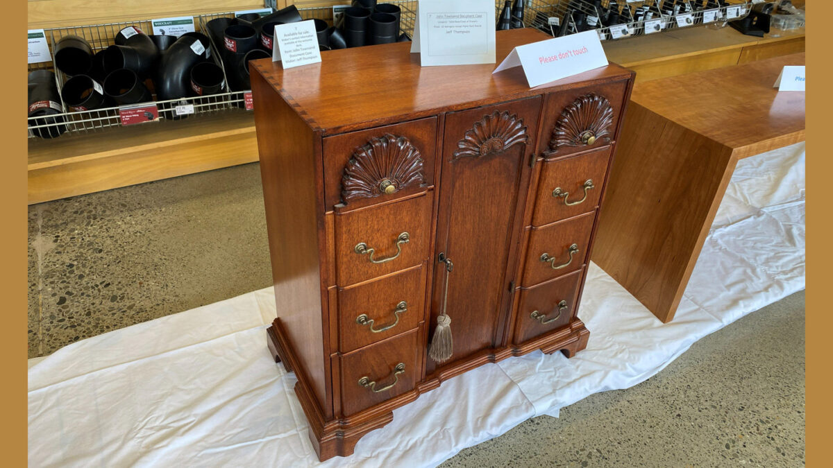 A wooden document case by Jeff Thompson featuring a central cabinet door flanked by three drawers on each side. The reddish-brown wood piece features carved shell motifs (fan carvings) above each drawer and the door, brass bail pulls, and a tassel hanging from the door handle. It stands on bracket feet, and fine dovetail joinery is visible along the top edge.
