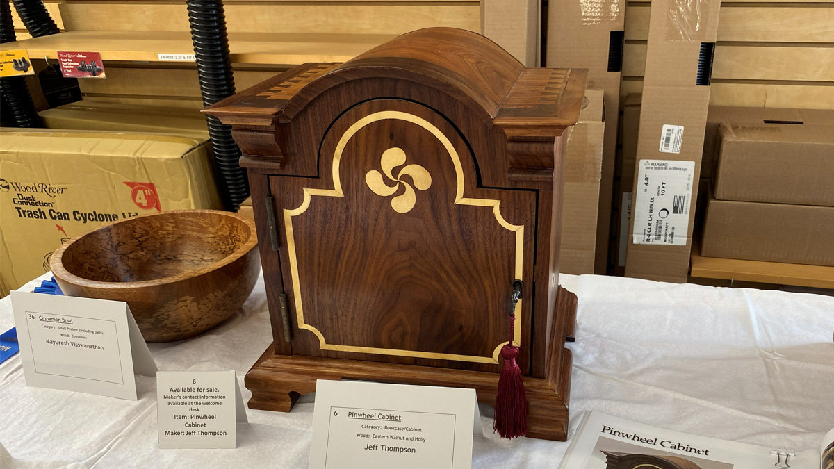 A dark wood cabinet with a curved top stands on a white tablecloth, featuring intricate light-colored wood inlay. The inlay creates an arch shape around the cabinet door and forms a four-bladed pinwheel design at the top center of the arch. A small metal latch with a red tassel hangs on the right side of the door. A small white display card in front of the items identifies the cabinet as the "Pinwheel Cabinet" made of Eastern Walnut and Holly by Jeff Thompson.