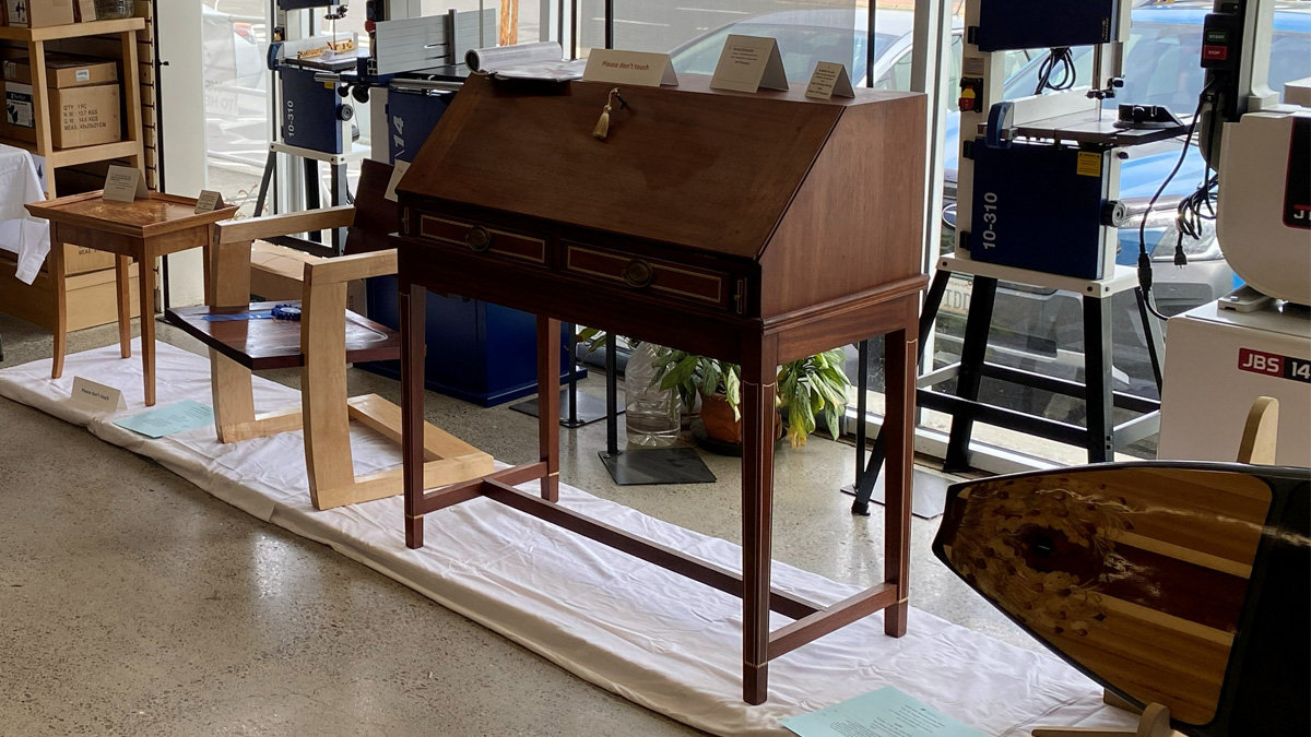 The Sheraton Writing Desk is a dark wood secretary cabinet with a slanted fall-front, featuring two drawers with geometric inlay accents and tapered legs connected by stretchers. Inspired by the book "Masterpiece Furniture Making" by Franklin Gottshall.