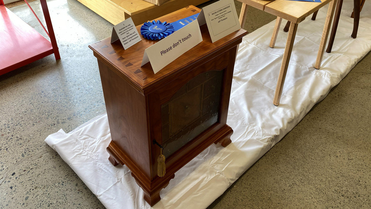 A small, reddish-brown wooden cabinet, identified by a card as a spice box made by Jeff Thompson. The box features a rich finish and a front door with a glass panel, and a gold tassel hangs from a latch on the side. The piece rests on a molded base with bracket feet. On top, there is a blue award ribbon and a card indicating it won in the "Bookcase/Cabinet" category.