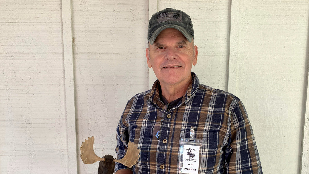 Jeff Wardwell stands facing forward, wearing a blue-and-brown plaid flannel shirt and a dark cap with an American flag patch. Clipped to his pocket is an Oakhurst Woodcarvers Rendezvous name badge. He holds up a wooden moose head carving with light-colored antlers, a project that he taught in his power carving class at the event.