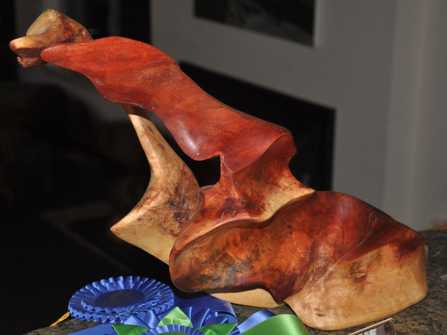 Abstract wooden sculpture by Jerry Gin featuring a dynamic, upward-curving form with polished reddish-brown and natural light wood tones, displayed next to blue and green award ribbons.