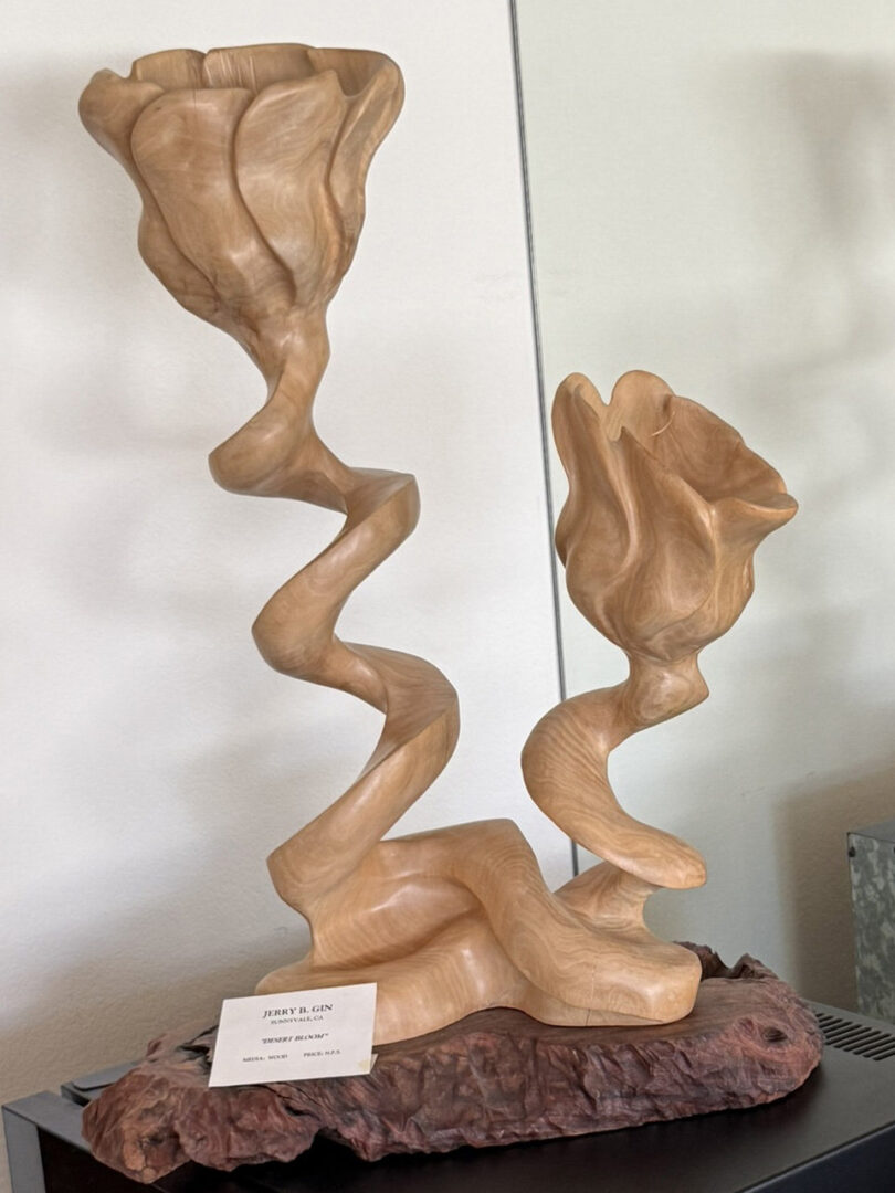 A wooden sculpture titled "Desert Bloom" by artist Jerry B. Gin, featuring two stylized flower blooms rising from twisting, helical stems mounted on a rough piece of bark.