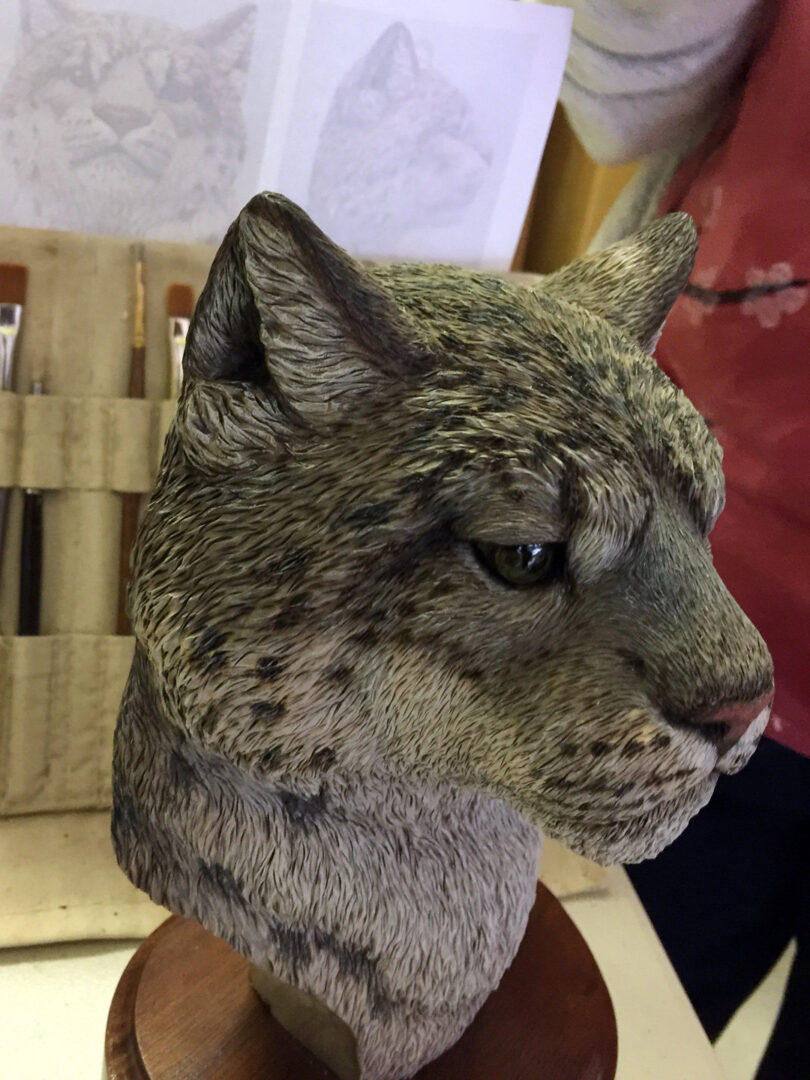This image displays a detailed wood carving of a mountain lion head and neck, mounted on a round wooden base. The sculpture features intricate texture simulating fur with fine lines, along with distinct dark spots on the cheek and jawline. The cougar has upright ears, a pink nose, and is looking slightly downward to the right. In the background, slightly out of focus, are artist's tools including a canvas roll holding paintbrushes and pencil reference sketches of a mountain lion face taped to a surface, indicating an art studio setting.