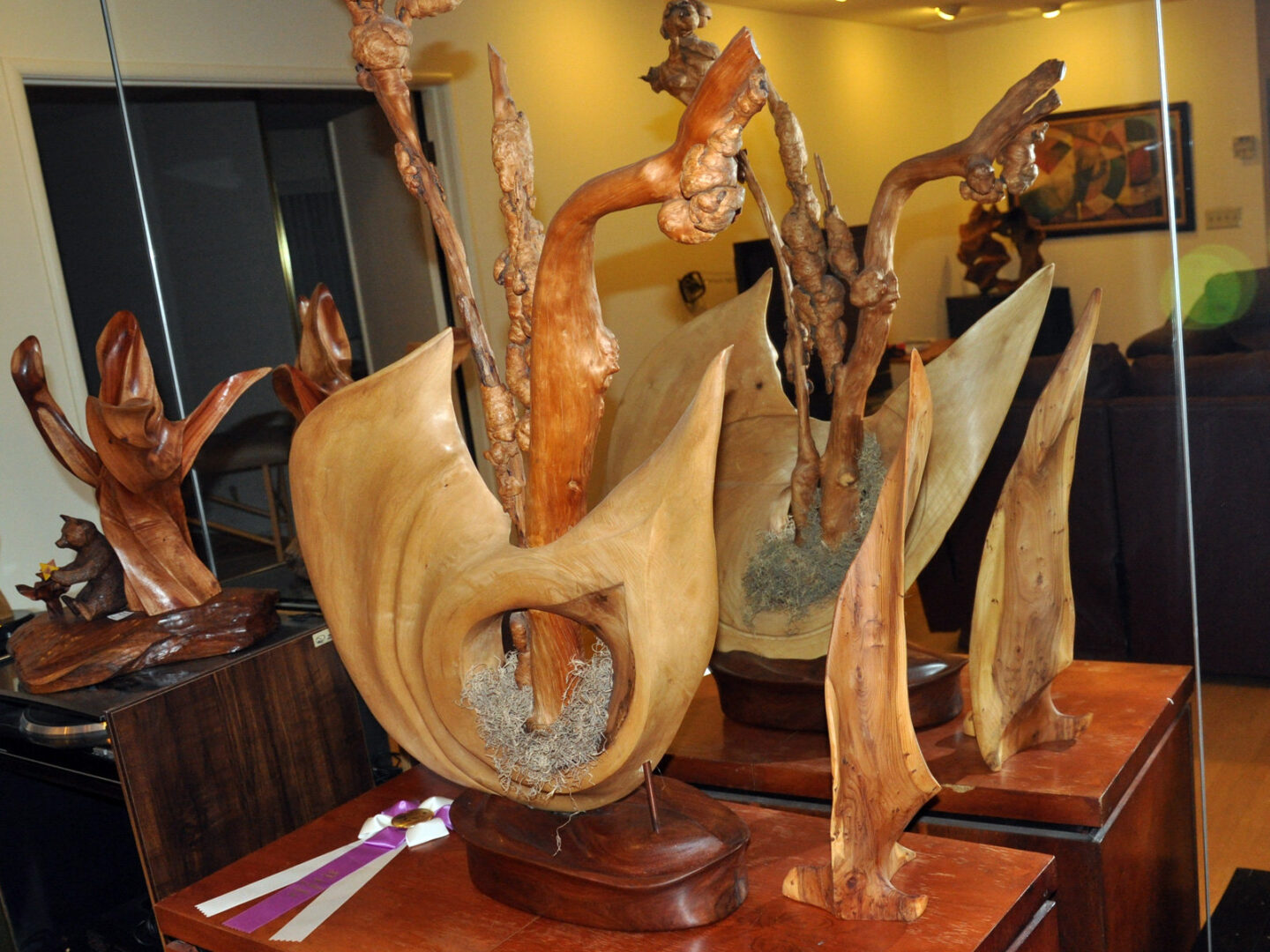 Swirling Form (2011), an abstract woodcarving by Jerry Gin, a member of Santa Clara Valley Carvers. The sculpture features smooth, curved hardwood with upward-reaching knotted branches and a central hollow containing fibrous material. A purple-and-white award ribbon sits at the base. The photograph captures both the original piece and its mirror reflection alongside another wooden carving visible to the left.