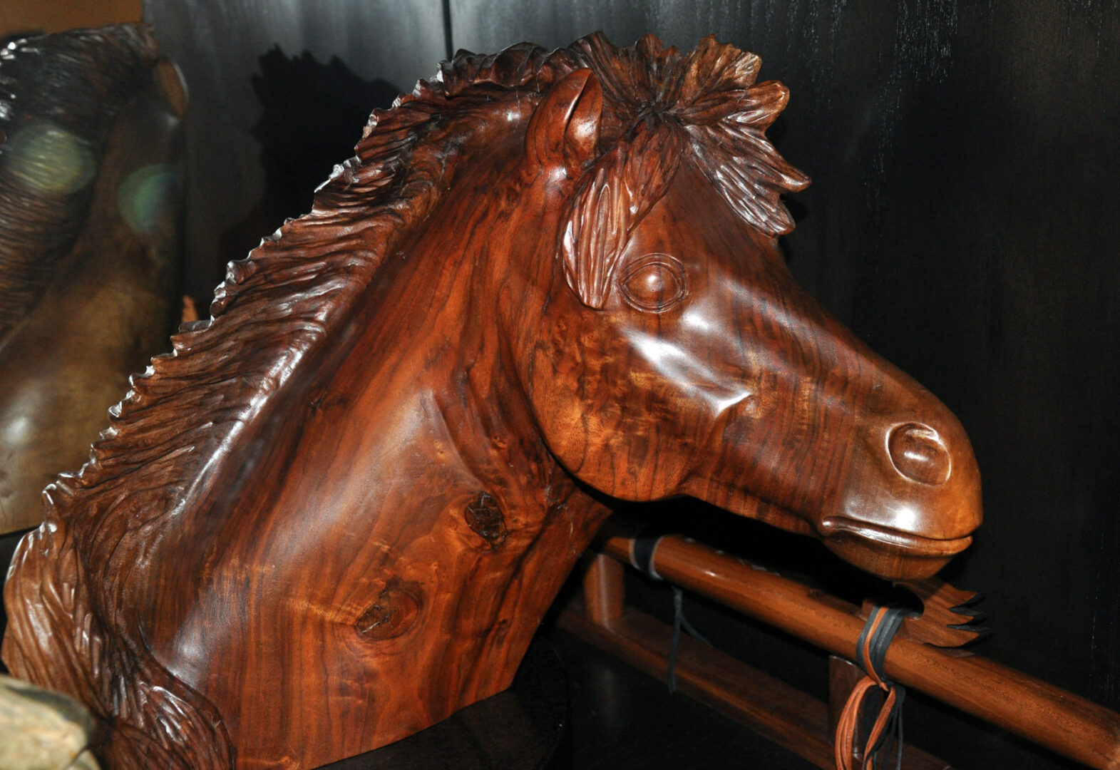 A close-up side view of a polished walnut wood carving of a horse's head and neck. The reddish-brown timber features prominent grain patterns and knots, with the mane carved in textured, backward-flowing strands. The sculpture rests on a wooden railing against a dark background. This piece is part of Jerry Gin's woodcarving gallery.