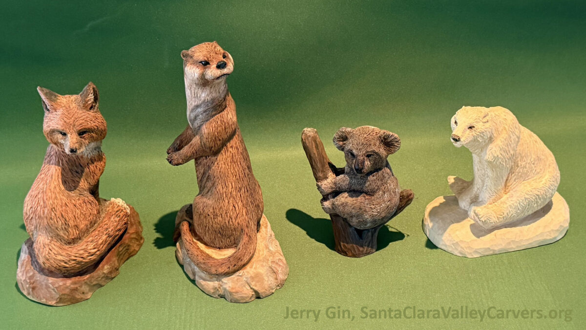 Four detailed wood carvings by artist Jerry Gin arranged on a green background: from left to right, a reddish-brown fox sitting on a rock, an upright otter standing on its hind legs, a small koala clinging to a piece of wood, and a white polar bear seated on an ice-like base.