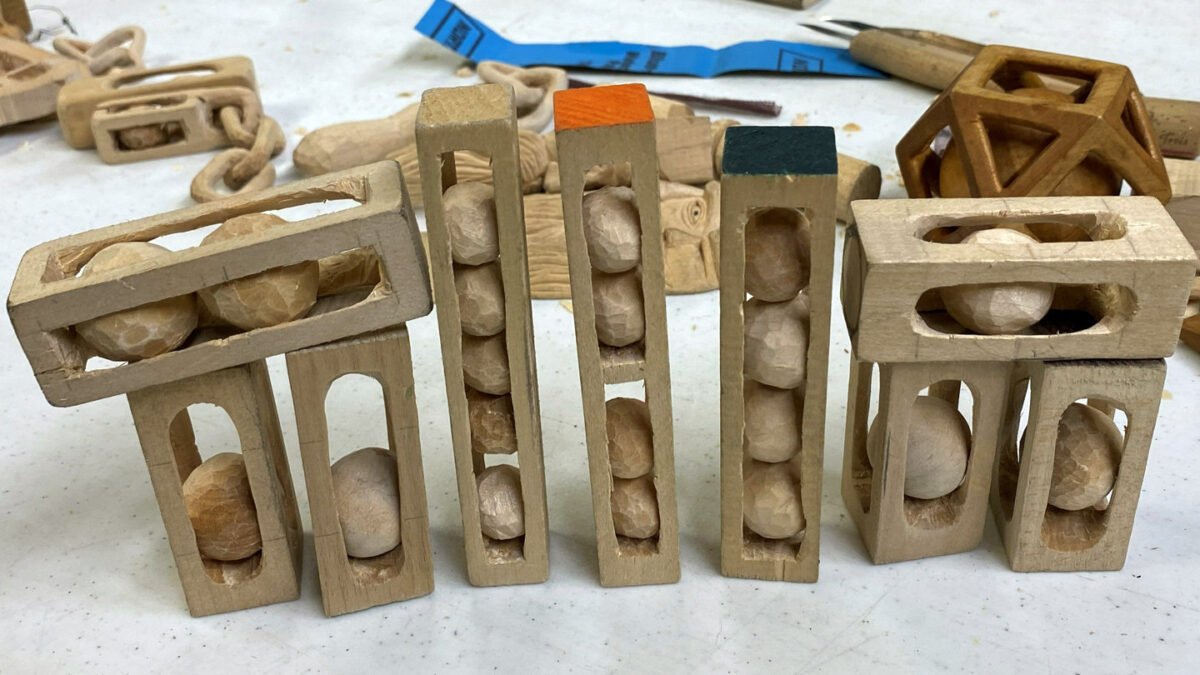A close-up view of hand-carved wooden "balls in cages" arranged on a white workbench at the 2026 Oakhurst Woodcarvers Rendezvous. The display features whittled basswood blocks hollowed out to hold rough-hewn spherical balls with visible knife marks. In the background, slightly out of focus, other projects like a wooden chain and a geometric lattice structure are visible.