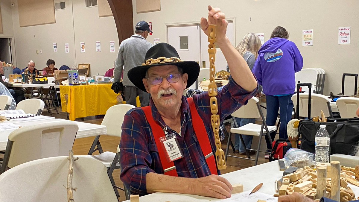 Jerry Schneider sits at a table inside a community hall, holding up a long, intricate wooden chain high with his left hand. He wears a black wide-brimmed hat adorned with a thick wooden chain band, red suspenders over a plaid shirt, and glasses. On the table in front of him are scattered wood shavings and small carved blocks, while other attendees and white folding tables fill the background.