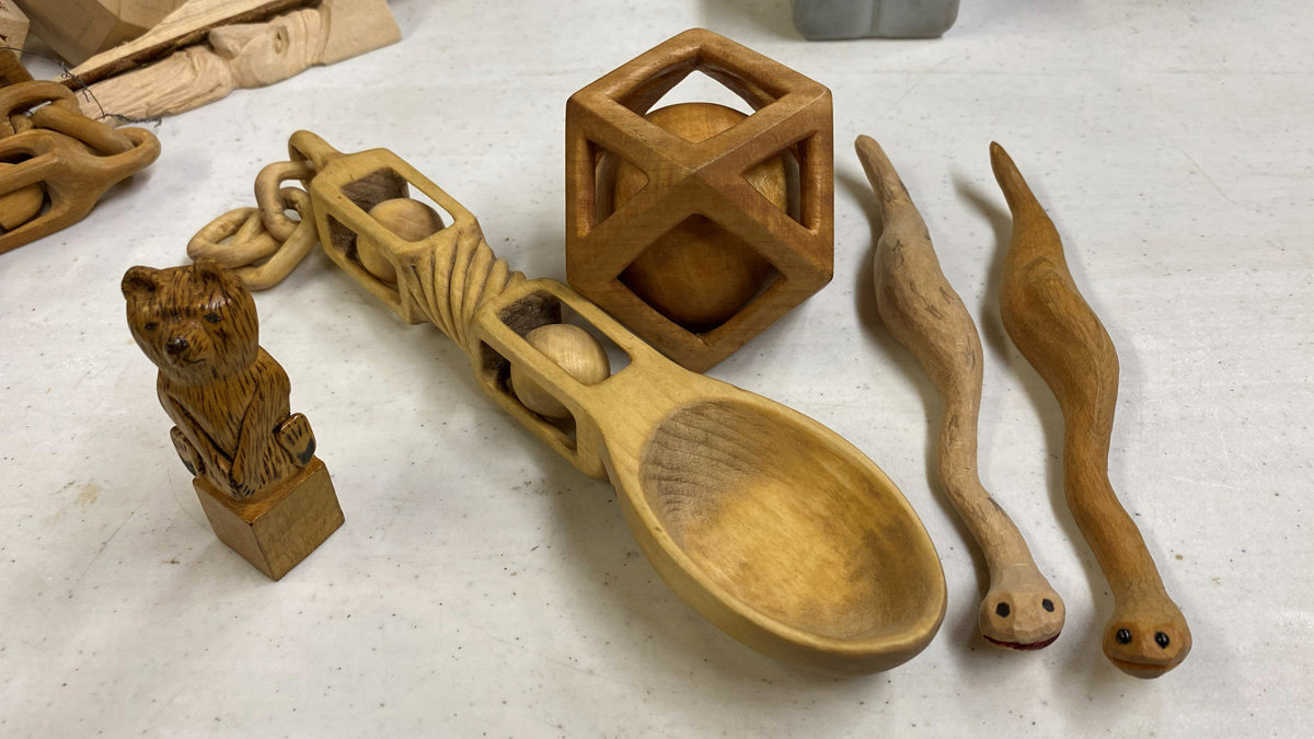 A collection of hand-carved wooden projects rests on a white surface, showcasing work by Jerry Schneider from the 2026 Oakhurst Woodcarvers Rendezvous. The arrangement includes a large wooden spoon with an intricate handle containing small balls, positioned in front of a cube-shaped ball-in-a-cage puzzle. To the right lie two slender, wavy wooden snakes with burned eyes, while a small detailed bear carving stands on a block to the left near a segment of a carved wooden chain.