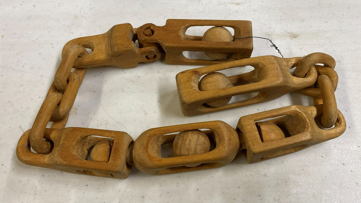 An intricate hand-carved wooden chain rests on a white speckled surface. The links are unique hollowed blocks containing smooth round spheres inside, connected by traditional oval rings. This piece demonstrates 'balls-in-cages' whittling techniques demonstrated by Jerry Schneider at the 2026 Oakhurst Woodcarvers Rendezvous.