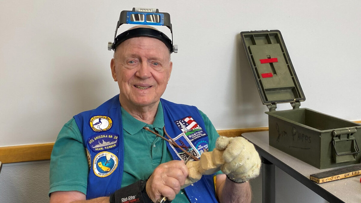 Member Jim Phipps is smiling while carving a piece of basswood with a folding carving knife, wearing magnifiers on his forehead and a protective glove. An open green ammunition box labeled with his name sits on the table to his right.