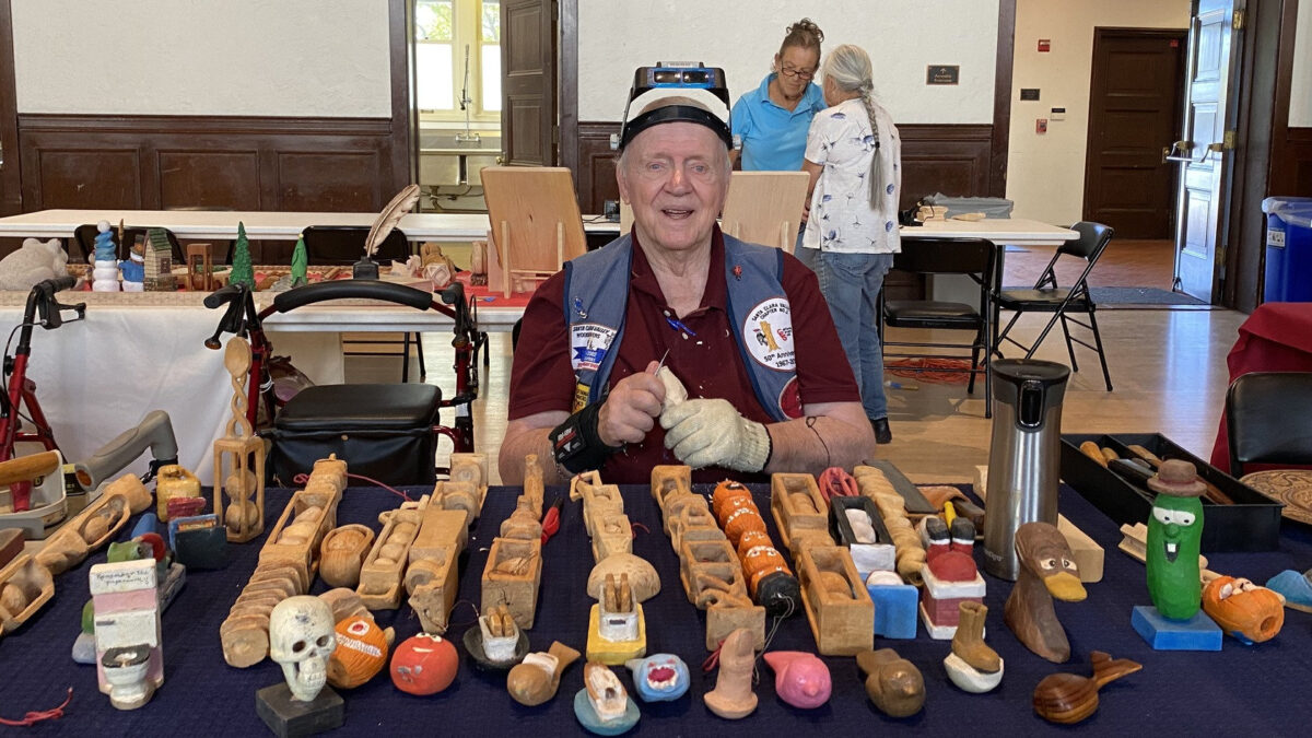Jim Phipps holds a carving knife and displays his whittling projects, including a carved pickle and skull, at the 2026 Tri-Valley Carvers Show in Pleasanton, California.