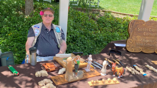 Jim Phipps at the table full of wood carvings. A huge Santa Clara Valley Carvers sign is visible on the right side of the picture.