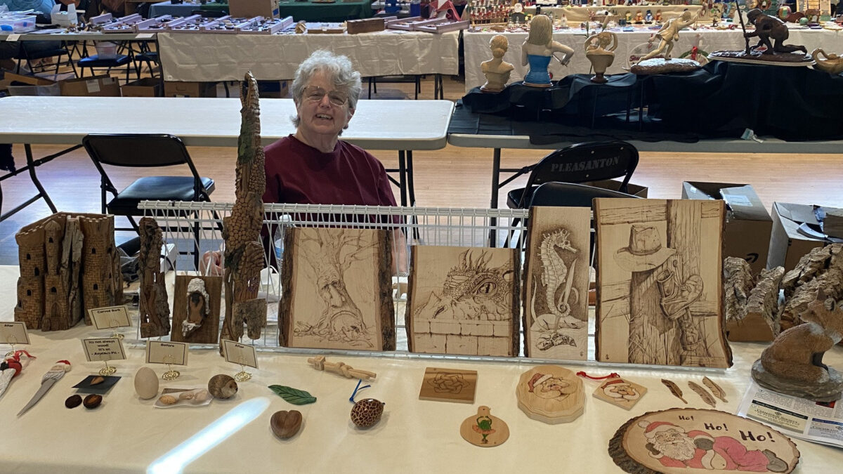 Joanne Carroll smiles behind her display table of woodcarving and pyrography art at the Tri-Valley Carvers Show, featuring intricate burned boards and textured bark carvings.