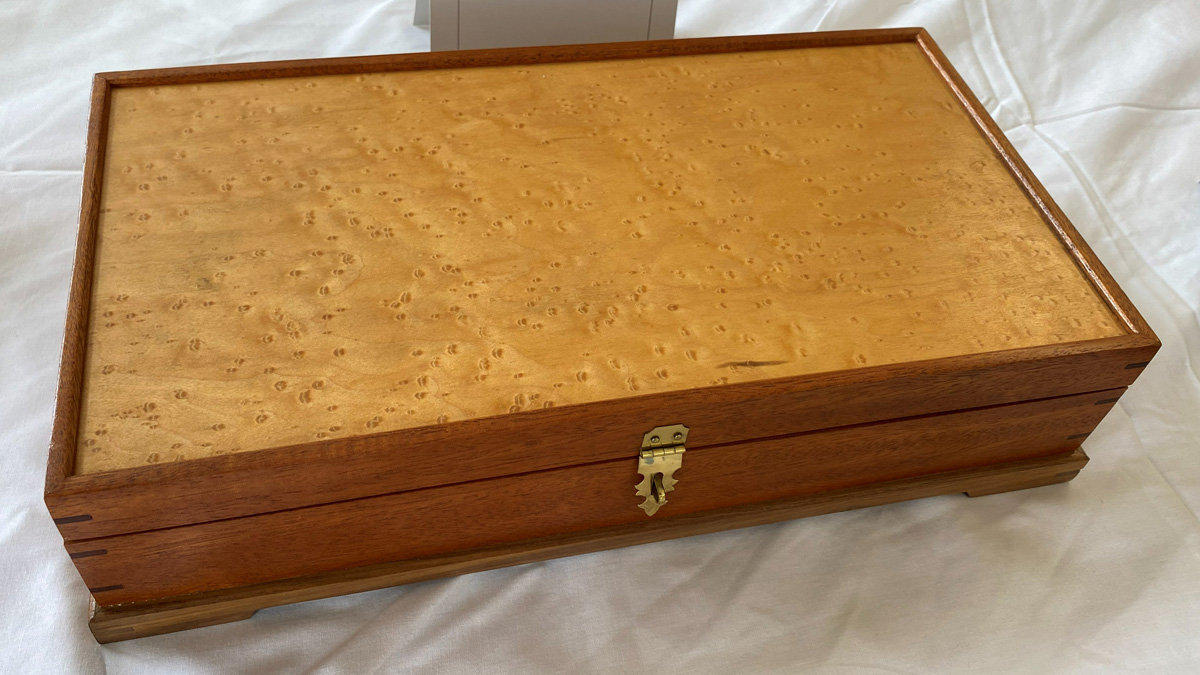 A rectangular wooden box featuring a lid made of light-colored birdseye maple with a distinctive swirling grain pattern, framed by a darker wood. The sides of the box are a medium reddish-brown wood with visible finger joints at the corners. A small brass latch is mounted on the front, and the box sits on a slightly recessed base.