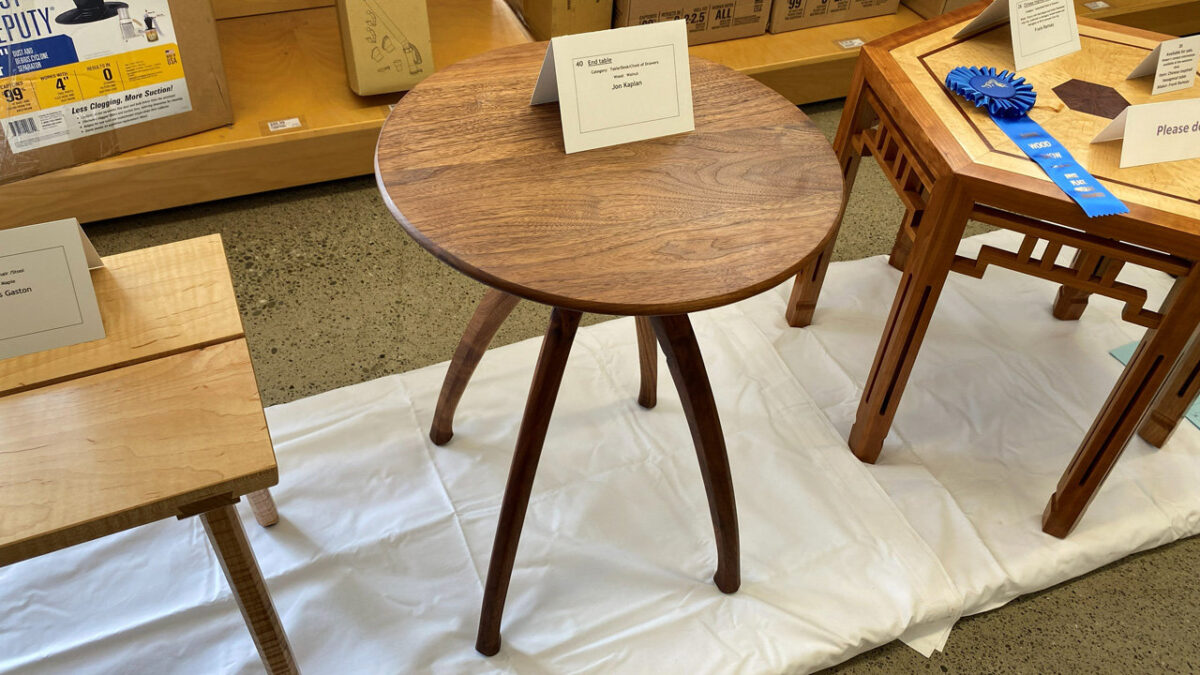 A round, three-legged wooden end table features a smooth, medium-brown top with visible grain and dark, curved legs that splay outward. A white card on the table identifies the piece as an "End table" by maker Jon Kaplan.