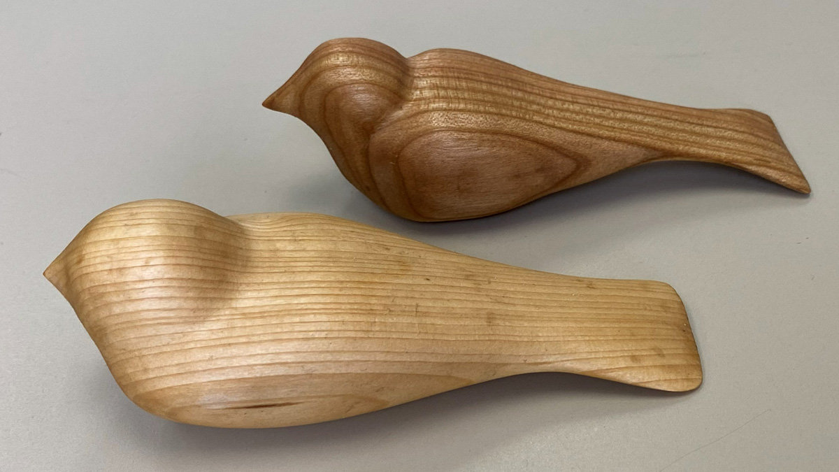 Two smooth, hand-carved wooden "comfort birds" created by Jonathan Jackel rest on a plain grey surface. The bird in the background is crafted from medium-brown wood with swirling grain patterns that suggest wings and tail feathers. In the foreground, a larger bird is carved from pale, light-colored wood with distinct straight horizontal grain lines running along its body. Both stylized figures are rounded and lack detailed facial features, facing toward the left.