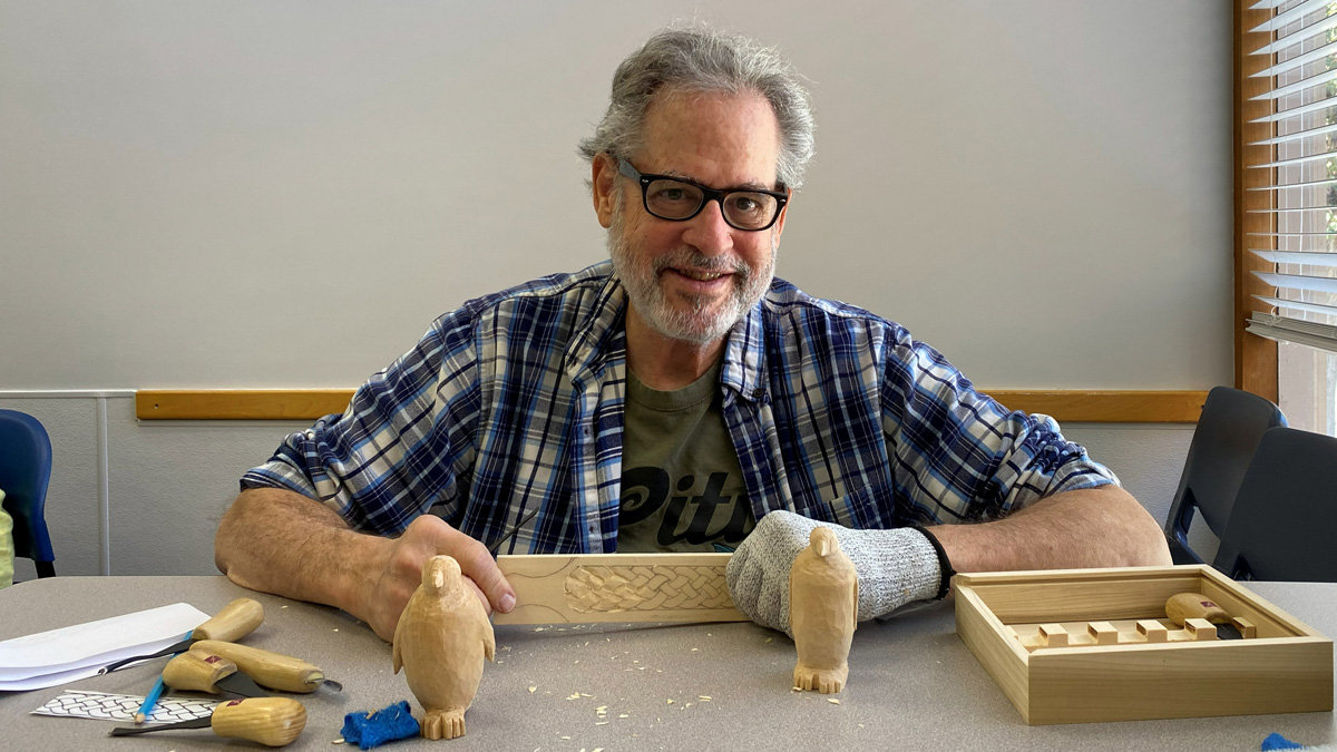 A smiling man (Jonathan Jackel) with gray hair and glasses sits at a work table covered in wood shavings. He wears a blue plaid shirt over a green T-shirt and has a thick protective glove on his left hand. He is making a leather strop by carving an intricate, braided pattern on a long, rectangular piece of light-colored wood (basswood). On the table in front of him stand two small, whittled penguin figurines. To his left are several carving tools with light handles and a sketch, while to his right sits a wooden box with woodcarving gouges.