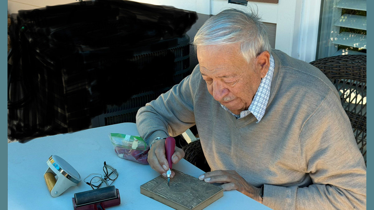 A man with white hair, Jordan Makower, focuses intently as he carves a design into a block of basswood with a red-handled tool.