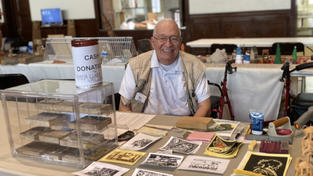 Jordan Makower smiles behind a table displaying his linoleum block prints, carving tools, and carved blocks at the 2026 Tri-Valley Carvers Show in Pleasanton, California.