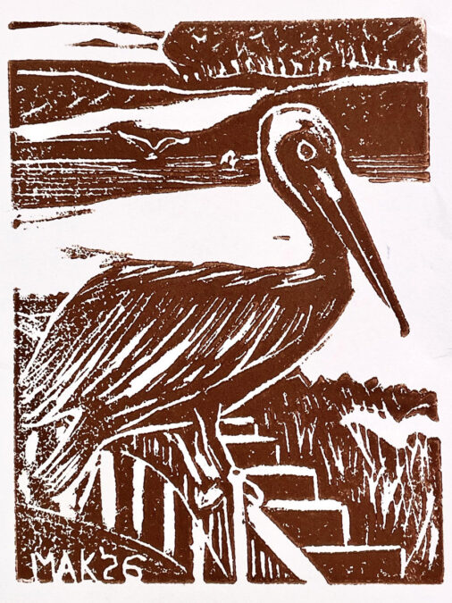A brown monochromatic linocut print featuring a large pelican standing in profile facing right. The bird is detailed with white carved lines representing feathers and a long beak. In the background, stylized horizontal bands depict water, a distant shoreline with trees, and a small flying bird. Stylized reeds or grass appear in the foreground near the bottom right. The signature "MAK '26" is visible in the bottom left corner.