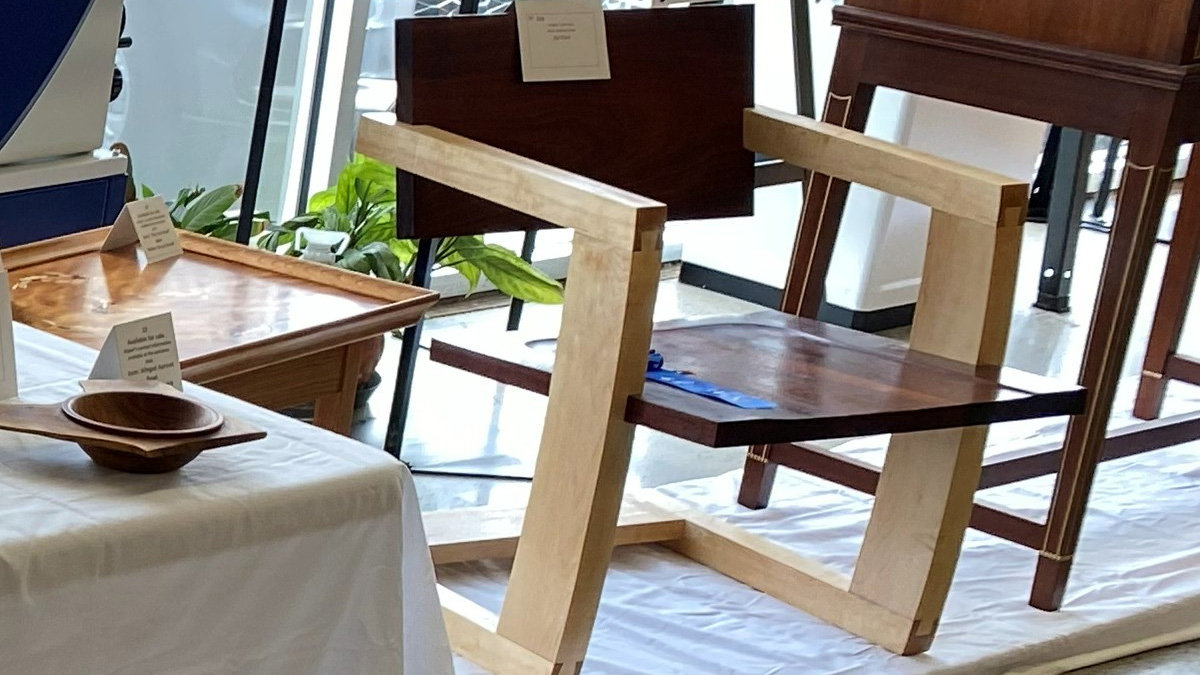 A modern armchair with a high-contrast design stands centrally, featuring a heavy, angular frame made of light-colored wood for the legs and armrests. The seat and tall rectangular backrest are crafted from dark wood and inspired by J. Rusten's Palo Alto Chair. A blue ribbon lies on the dark wooden seat, indicating an award.