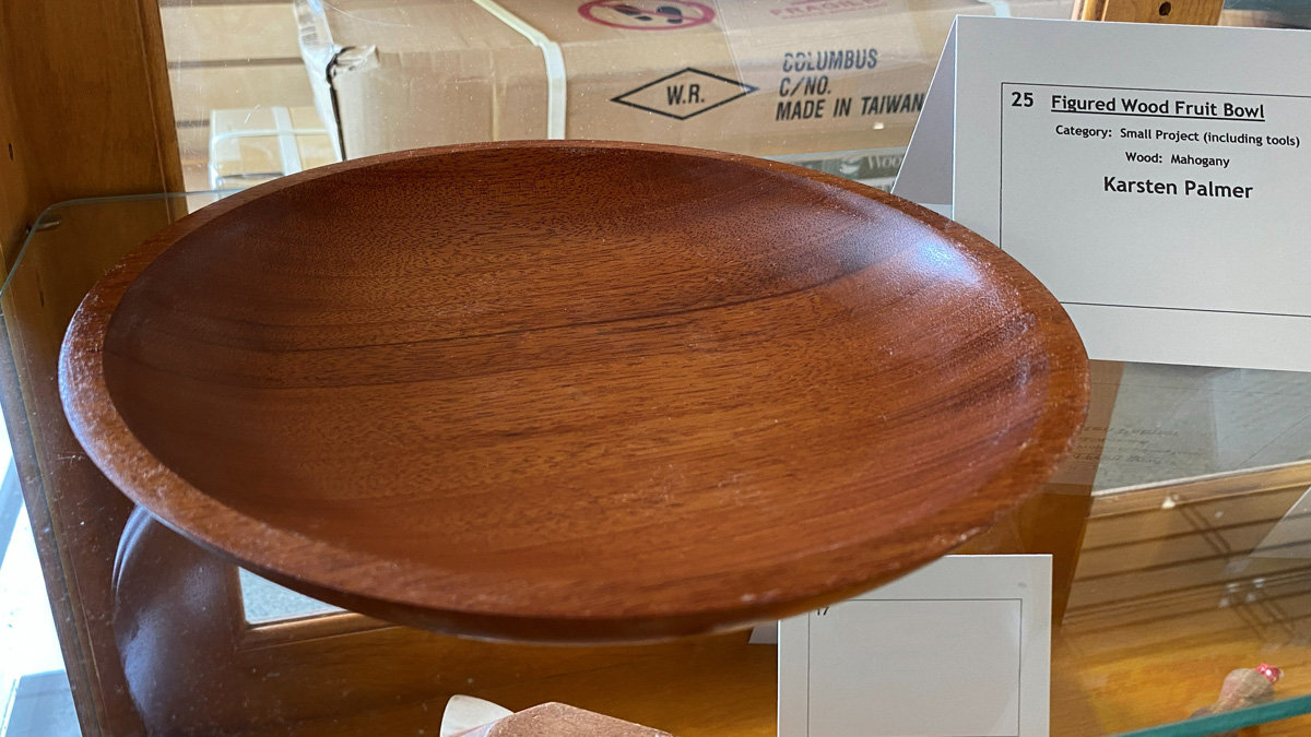 A polished, reddish-brown wooden bowl or platter sitting on a glass shelf. The wood, identified by a nearby label as mahogany, has a smooth finish with visible grain patterns. A white card next to the object identifies it as a "Figured Wood Fruit Bowl" turned by Karsten Palmer.