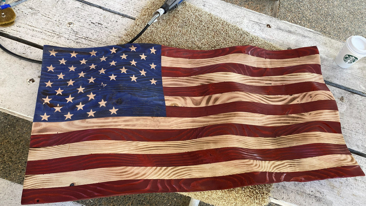 A large wooden relief carving of an American flag resting on a workbench covered in sawdust. The artwork features a wavy, undulating surface texture designed to make the flag look like it is waving in the wind. The stripes alternate between natural light-colored wood and a deep reddish-brown stain, while the blue canton is stained a deep shade, with stars carved out of the lighter natural wood. A rotary carving tool is visible at the top edge of the frame near the carving.