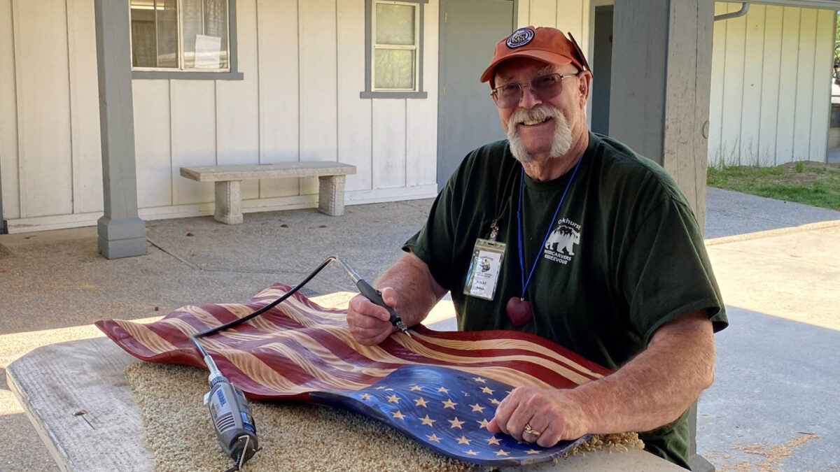 A man, Kelly Hall, with a white goatee and glasses, wearing an orange baseball cap and a dark green T-shirt with "Oakhurst Woodcarvers Rendezvous" printed on it, smiles as he demonstrates woodcarving techniques. He is seated at a table covered in sawdust, holding a rotary carving tool over a carved wooden American flag that features red and natural-wood stripes and a blue field with gold stars.