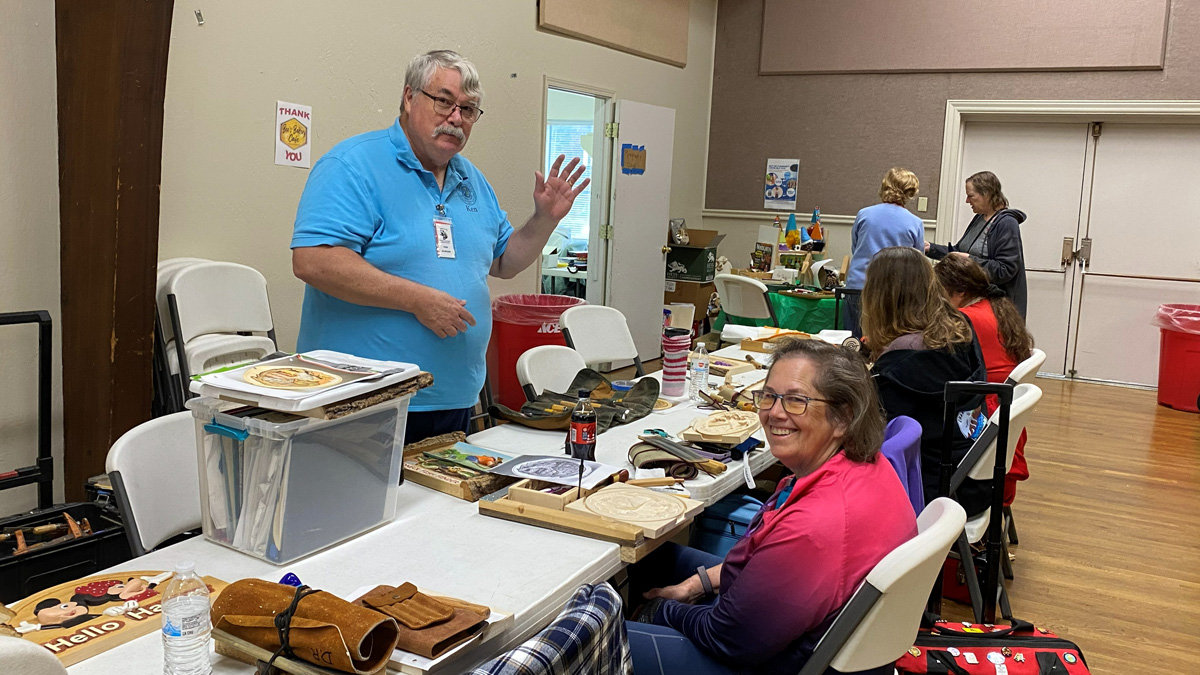 In a workshop setting at the Oakhurst Woodcarvers Rendezvous, instructor Ken Morgan stands behind a table filled with woodcarving supplies and blocks of basswood, gesturing as he teaches a relief carving class. A smiling student in a pink shirt sits in the foreground near rolled leather tool pouches, while other attendees work along the tables in the background.