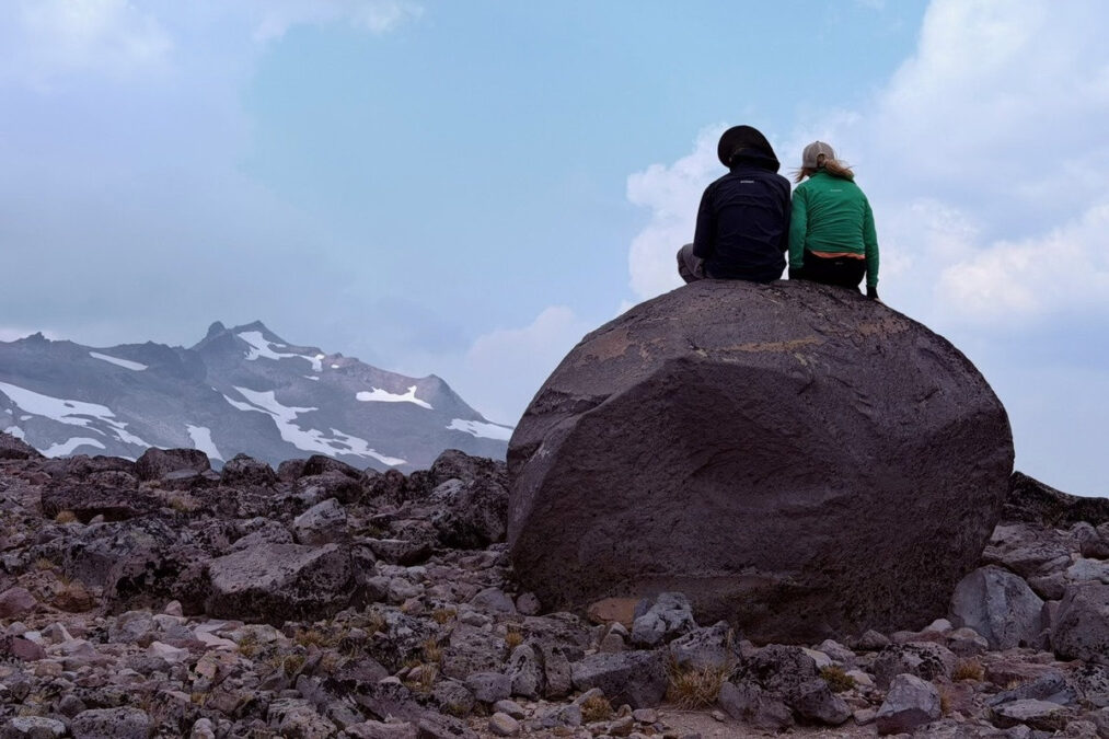 Two people, seen from behind, sit atop a massive boulder, overlooking a glacial landscape with snow-dusted mountains in Northern Washington State. This is a reference photo for the Kids & Perseids block print project.