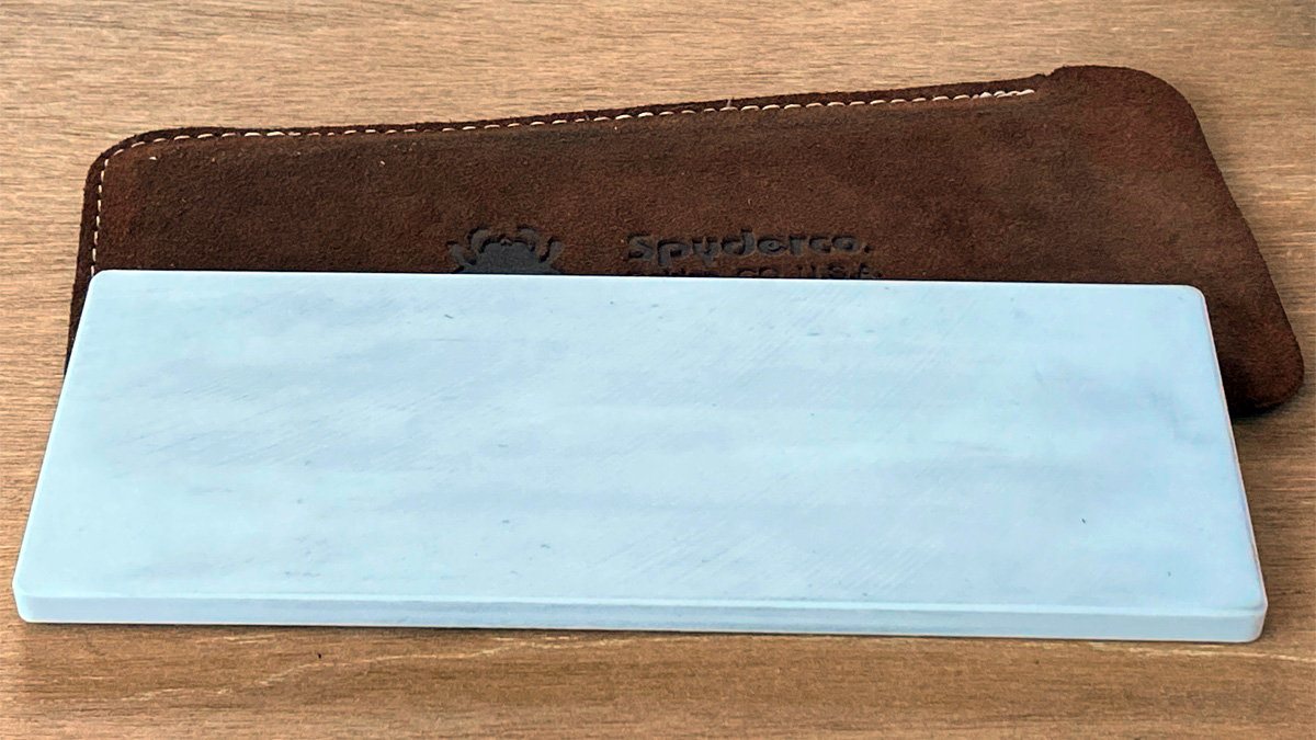 A large, rectangular, white ceramic sharpening stone rests on a wooden surface in front of a brown suede storage pouch embossed with the Spyderco logo and text.