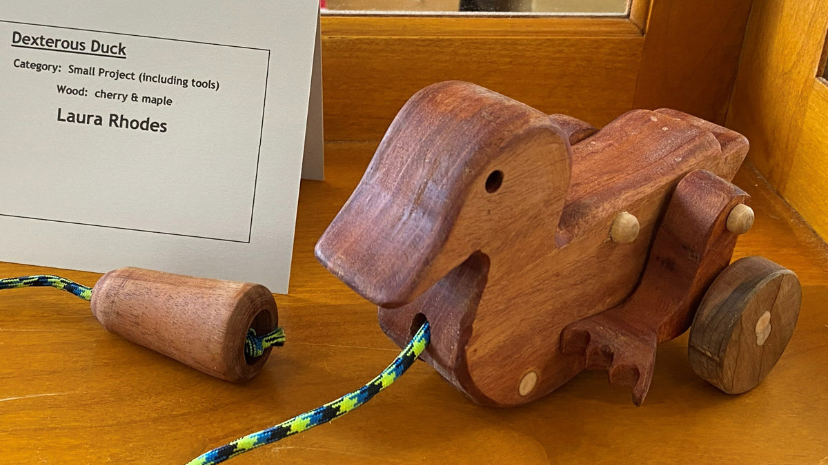 A wooden pull-toy duck made primarily of reddish-brown cherry wood with lighter maple accents sits on a wooden surface. The duck features a large, flat beak, a hole for an eye, and wooden wheels attached to its body. A blue, yellow, and black braided rope is threaded through a hole in the front of the duck, leading to a small wooden handle resting on the shelf. To the left of the toy, a white information card identifies the piece as the "Dexterous Duck" by Laura Rhodes, noting it is a "Small Project" made of cherry and maple wood.