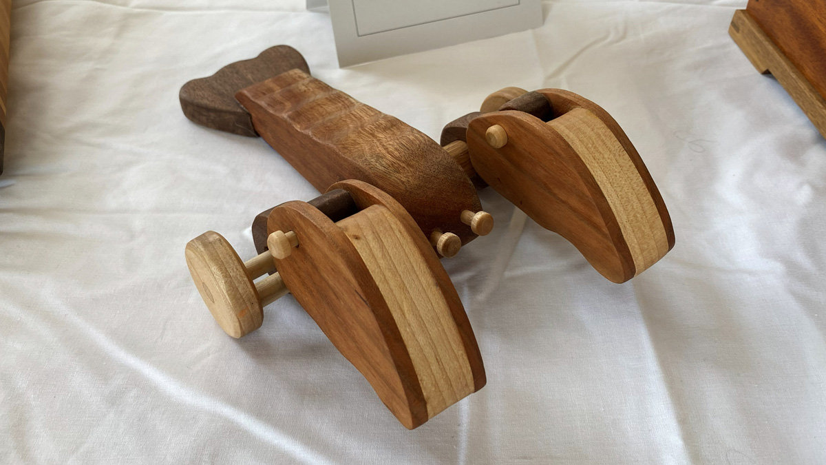A handcrafted wooden toy lobster, identified as a "Loco Lobster," is displayed, showcasing intricate woodworking. The toy features a central body made of reddish-brown wood with a textured, segmented appearance, attached to a dark, fan-shaped tail fin at the rear. Large, curved side pieces resembling lobster claws are constructed from laminated wood with light and dark tones. These side pieces are mounted on wooden axles equipped with small, light-colored wooden wheels, indicating the toy is designed to roll. Small wooden dowels protrude from the front of the body, serving as antennae. The piece highlights the natural grain and contrast of the different wood species used.